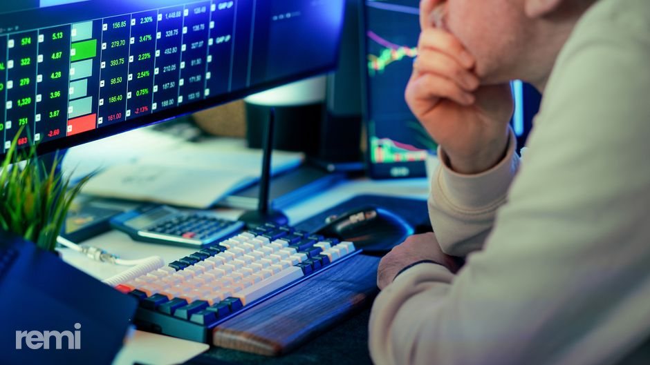 An investor sits at trading screens, hand on chin, watching red and green market data — the tension of waiting out a downturn